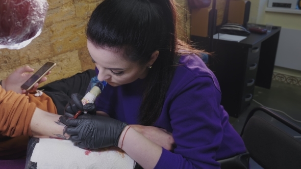 Brunette Female Tattooer in Violet Clothing Making Tattoo on the Man's Arm of Her Client with Gun in