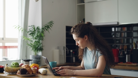 Young Concentrated Cheerful Woman Working in Kitchen Typing Laptop Computer During Breakfast