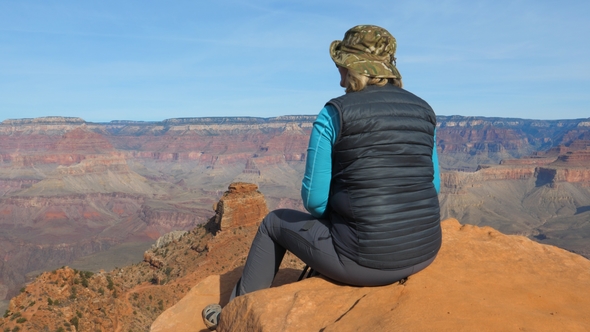 Woman Tourist Sitting On Rock Dangling Her Legs, And Admires Views Grand Canyon