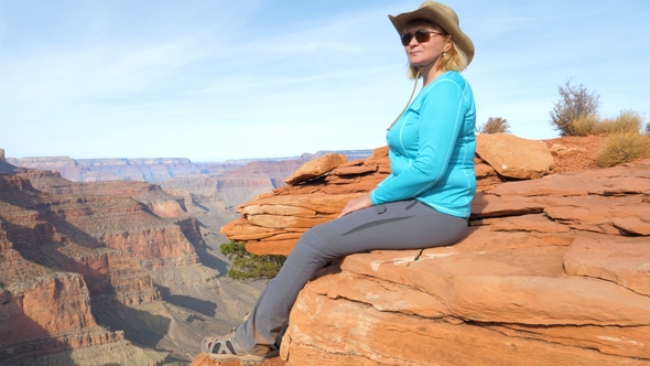 Woman Tourist Sitting On Rock Dangling Her Legs, And Admires Views Grand Canyon