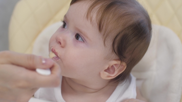 Baby Eats Porridge From Spoon, Spits and Smiles Sitting on Highchair in Kitchen.