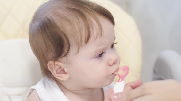 Baby Eats Porridge From Spoon, Spits and Smiles Sitting on Highchair in Kitchen