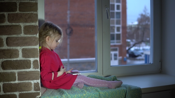 A Little Sweet Girl in a Pink Dress Sits on the Windowsill and Uses a Smartphone