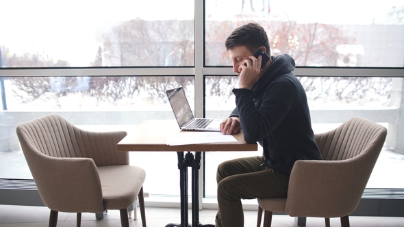 a Young Businessman Talking on the Phone and Working at a Laptop in a Cafe