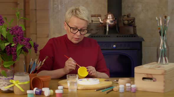 Focused Artist Paints Surface of Yellow Pot with Paintbrush on Wooden Table