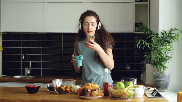 Attractive Happy Girl Dancing in Kitchen While Browsing Social Media on Smartphone and Listening To