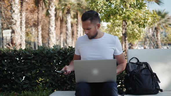 Smiling Young Man Working on the Laptop Sitting in Bench with Travel Case and Bag Backpack Tourist