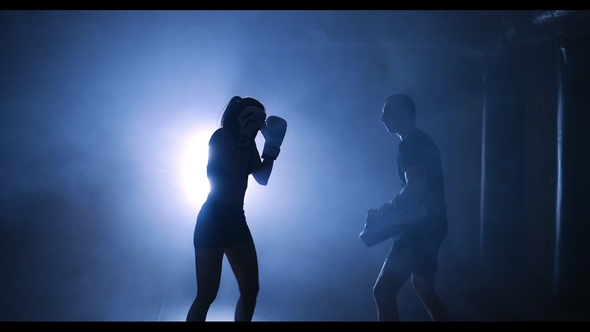 Silhouettes of a Female Boxer Punching a Boxing Bag with Boxing Gloves in a Smoky Gym