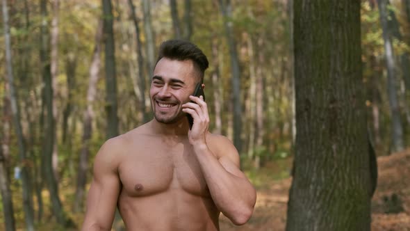 Strong Guy With Naked Torso Stands In Autumn Park And Speaks On Mobile Phone.