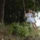 of a Charming Little Girl in a White Embroidered Dress Swaying on a Wooden Swing Seat in a Garden - VideoHive Item for Sale