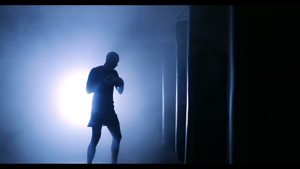 Strong Male Boxer Punching a Boxing Bag with Boxing Gloves in a Smoky Gym