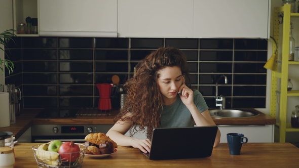 Young Curly Pretty Serious Caucasian Woman Working on Laptop and Talking on Phone Sitting in Modern