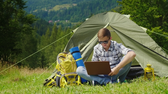 A Young Male Tourist Uses a Laptop in a Camping Near a Tent