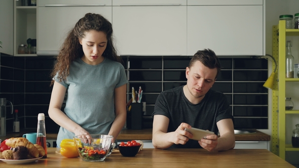 Attractive Couple in the Kitchen Man Playing Video Game on Smartphone While His Girlfriend Cooking