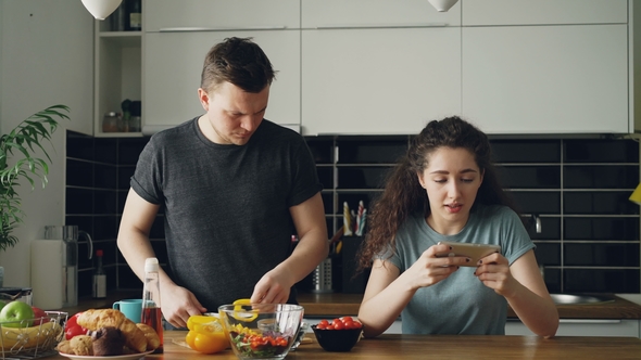 Attractive Couple in the Kitchen at Home. Curly Girl Playing Video Game on Smartphone.