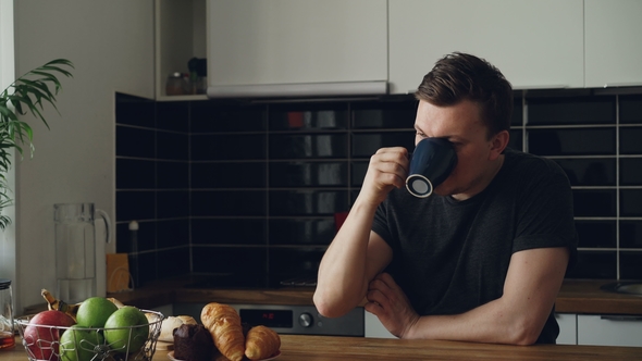 Portrait of Young Happy Caucasian Man Sitting at Table in Modern Spacious Kitchen, Looking Through