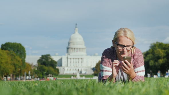 A Woman with a Smartphone Lies on a Lawn Against the Background of the Capitol in Washington