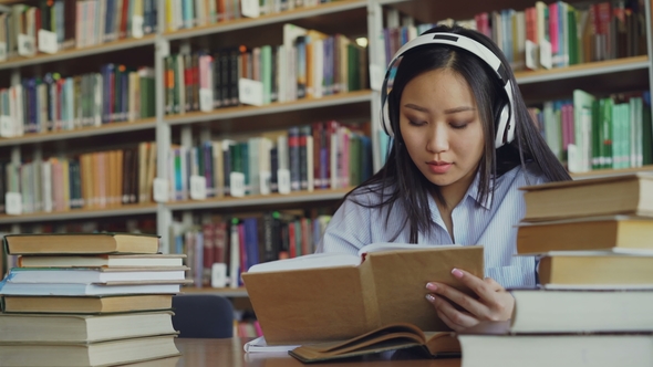 Pretty Positive Asian Teenage Student with Headphones on Head Listening To Music Is Sitting at Table