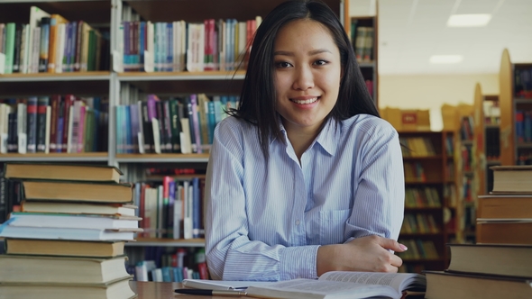 Portrait of Young Beautiful Asian Female Student Sitting at Table with Piles of Textbooks in Library
