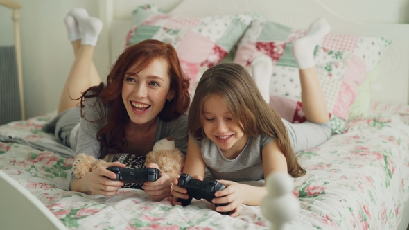 Happy Young Mother and Cute Cheerful Daughter Playing Computer Console Games on TV While Lying on