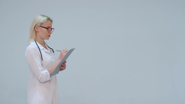 Blonde Doctor Writing on a Clipboard While Smiling Against a Grey Background