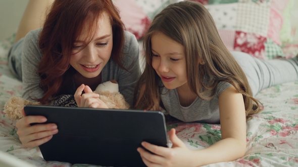 Smiling Cute Girl and Young Mother in Pajamas Laughing and Looking in Digital Tablet While Lying