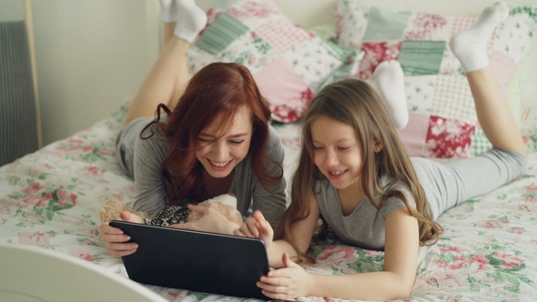 Beautiful Young Mother and Her Cute Daughter in Pajamas Laughing and Looking in Digital Tablet While