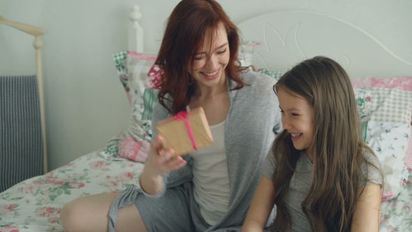 Young Happy Mother Giving Gift Box To Her Little Cute Daughter Celebrating Birthday Sitting on Bed