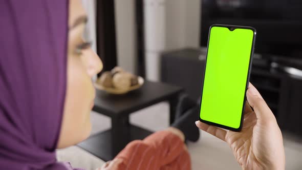 A Muslim Woman Looks at a Smartphone with Green Screen in an Apartment  Closeup From Behind