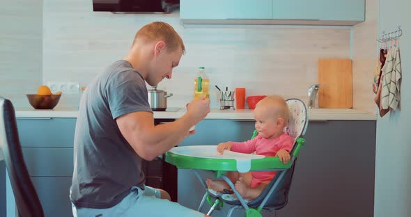 Handsome Young Man Sitting in the Kitchen and Feeding His Baby Porridge