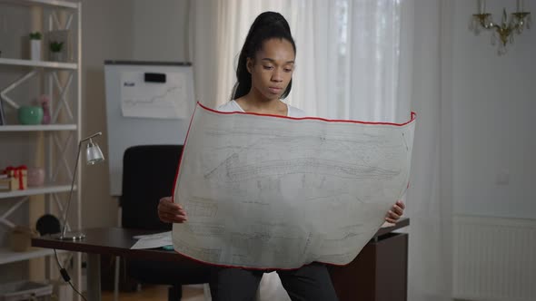 Portrait of Concentrated Skilled Female Architect Sitting on Table Opening Blueprint Examining Plan