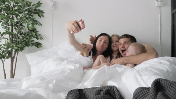 Cheerful Young Family Making Funny Selfie While Laying in Bed with Mobile Phones