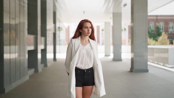 Beautiful business woman red hair in white coat stands near an office building