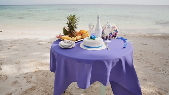 A Decorated Wedding Table with Fruits, Cake, Champagne and Wine Glasses on a Sandy Ocean Beach