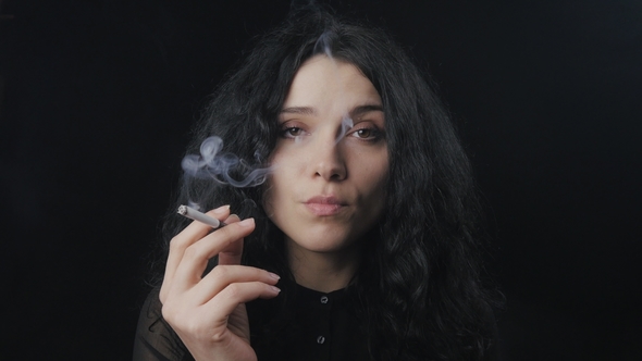 Young Brunette Woman with Curly Hair Is Smoking a Cigarette and Looking at the Camera on