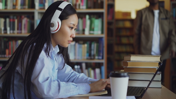 Beautiful Asian Female Student in Headphones Sitting at Desk in Library Working Concentrated at