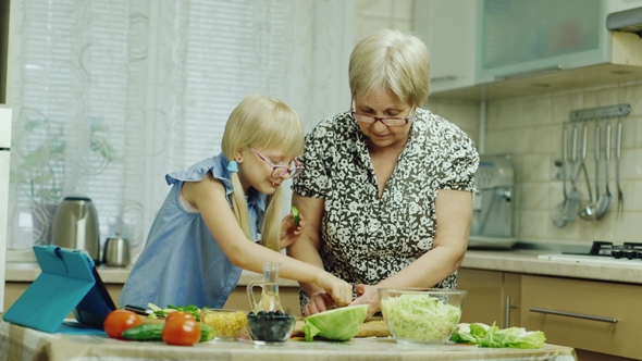 Grandmother Teaches Her Granddaughter To Prepare a Salad, Use a Tablet. Together Have a Good Time