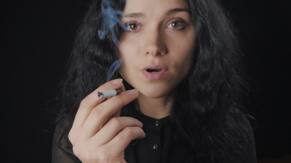 Portrait of Young Brunette Woman with Curly Hair is Smoking a Cigarette and Looking at the Camera on