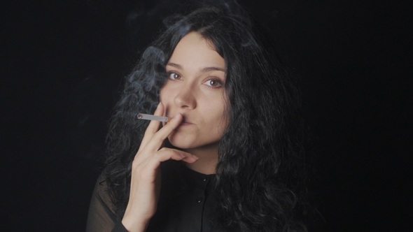 Portrait of Young Brunette Woman with Curly Hair Is Smoking a Cigarette and Looking at the Camera on