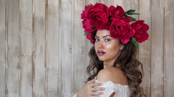 Girl Posing in Front of Camera. Young Woman in a Wreath of Scarlet Peonies on Her Head, Dark Long
