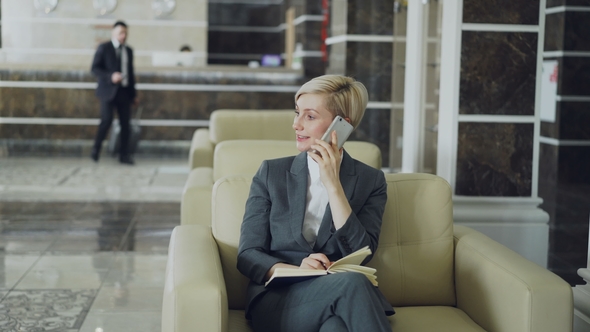 Attractive Businesswoman Sitting in Armchair Talking Mobile Phone with Notepad While Businessman