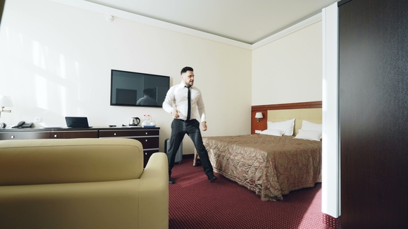 Businessman with Luggage Entering Hotel Room and Jumping on Bed Happily Then Lying Relaxed Smiling