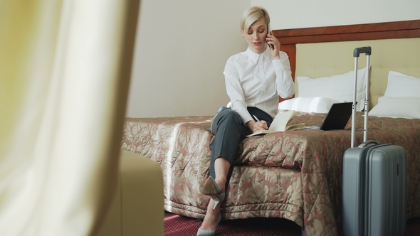 Cheerful Businesswoman Talking Mobile Phone and Writing Notes in Notepad While Sits on Bed in Hotel