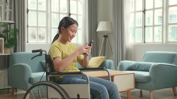 Asian Kid Girl Sitting In A Wheelchair While Using Smart Phone At Home