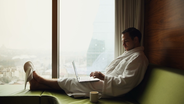 Young Businessman Working on Laptop Computer Wearing White Bath Robe Sitting near Window