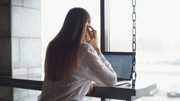 Young Businesswoman Dressed in White Shirt Is Sitting in Cafe at Wooden Table in Front of Laptop and