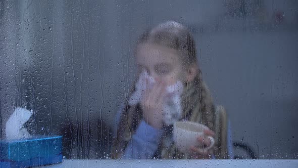 Little Girl Suffering From Flu Behind Rainy Window Sneezing and Drinking Hot Tea