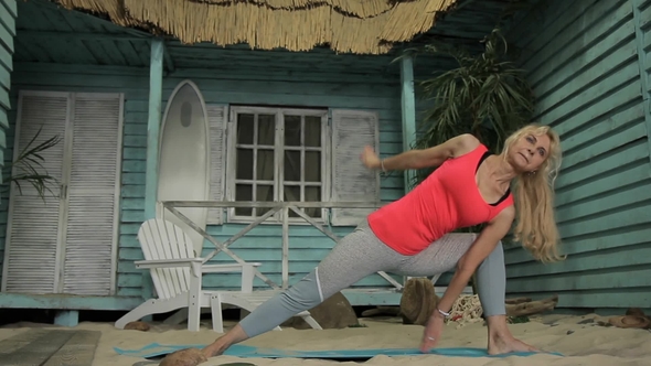 Mature Woman Is Having a Yoga Training Near the Blue Bungalow on Sandy Beach.