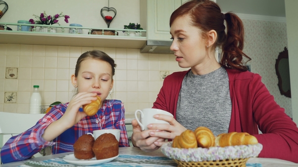 Cheerful Mother and Cute Daughter Having Breakfast Eating Muffins and Croissants Talking at Home in