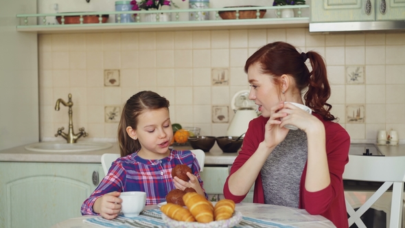Happy Mother and Cute Daughter Having Breakfast Eating Muffins and Talking at Home in Modern Kitchen
